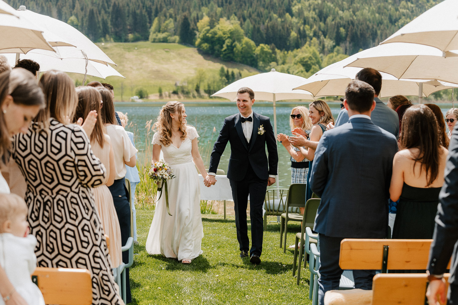 Das Brautpaar befindet sich am Badestrand des Neusacherhofs . Es hat gerade die Standesamtliche Trauung stattgefunden und sie lachen in die Kamera und ziehen zwischen den Gästen aus. Hochzeitsfotograf Valentino Zippo, Heiraten in Kärnten, Hochzeit am See in Kärnten, Hochzeitslocation am See, Hochzeitslocation Weissensee, Hochzeitsfotograf Österreich
