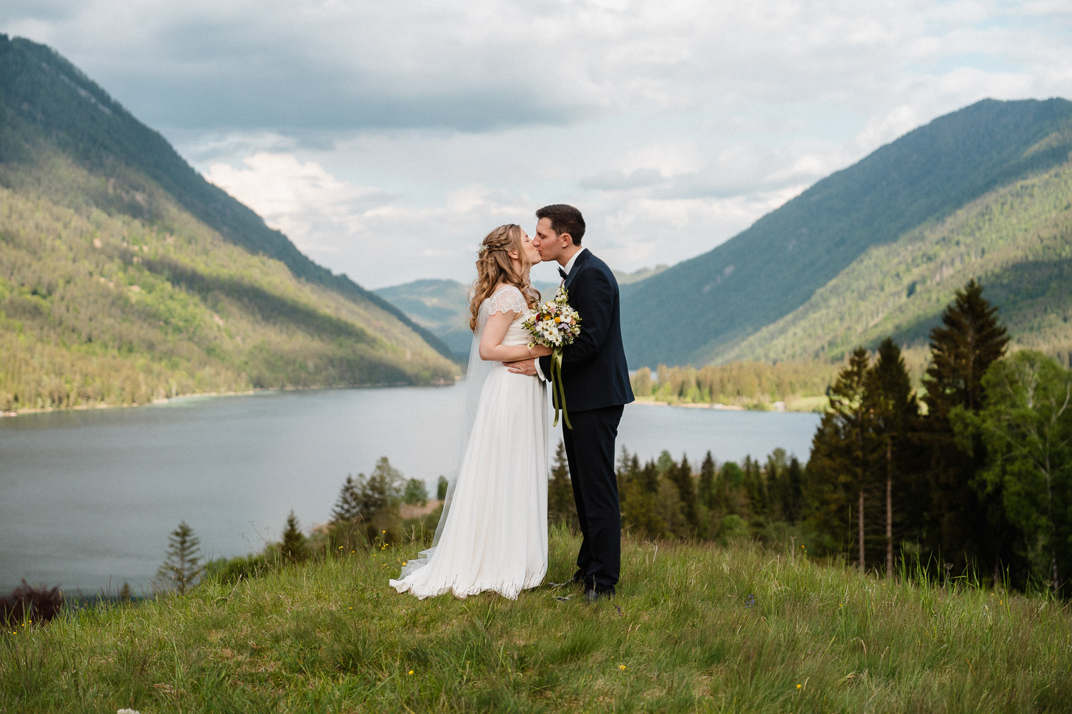 Hochzeitsfoto am Weissensee in Kärnten. Das Brautpaar steht auf einer Wiese über dem See und küsst sich. Aufgenommen von Hochzeitsfotograf Valentino Zippo aus Kärnten. Hochzeit am Weissensee, Hochzeitslocation, Hochzeitsfotos