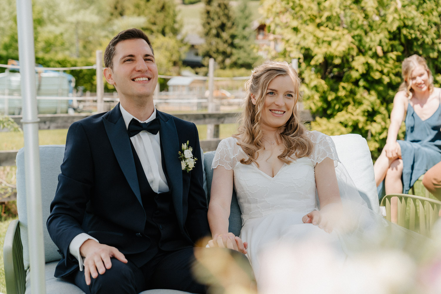Das Brautpaar sitz am Badestrand des Neusacherhofs auf ihrer Bank. Es findet gerade die Standesamtliche Trauung statt und sie lachen in die Kamera. Hochzeitsfotograf Valentino Zippo, Heiraten in Kärnten, Hochzeit am See in Kärnten, Hochzeitslocation am See, Hochzeitslocation Weissensee, Hochzeitsfotograf Österreich