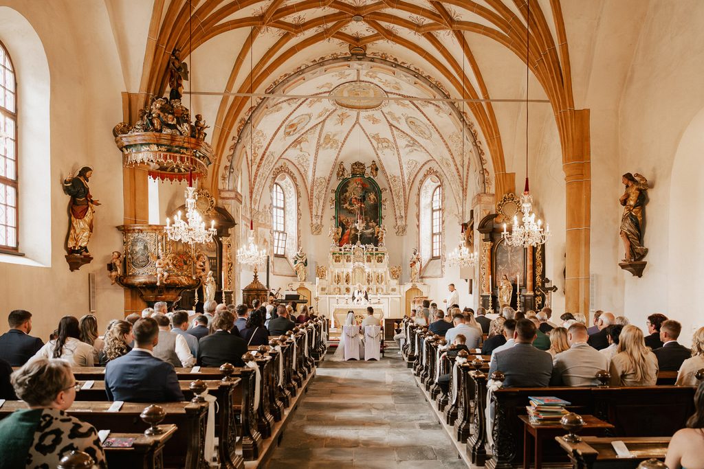 Weite Innenaufnahme der Kirche in Lieseregg in Seeboden am Millstättersee bei einer Hochzeit mit dem sitzenden Brautpaar. Hochzeit am Millstättersee, Kirche, Heiraten am Millstättersee, Hochzeitsfotograf Millstättersee, Hochzeitsfotograf Kärnten, Kirchliche Trauung