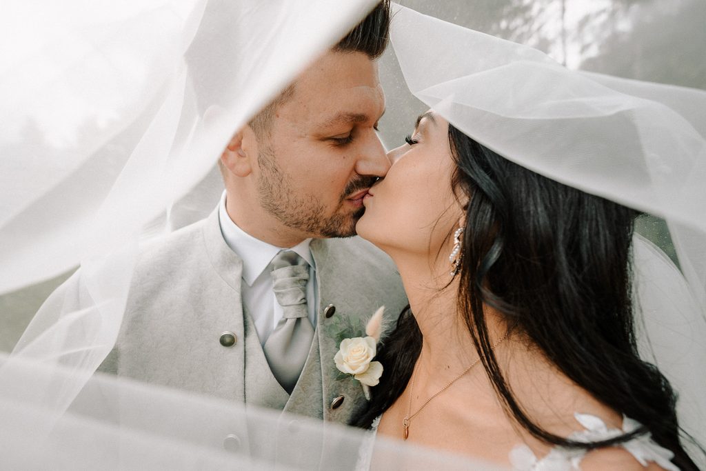 Das Hochzeitspaar in Seeboden am Millstättersee küsst sich unter dem Schleier vor der Kirche in Lieseregg. Hochzeitsfotograf Seeboden am Millstättersee, Hochzeit Millstättersee, Hochzeitsfotos, weddingphotographer, Millstatt