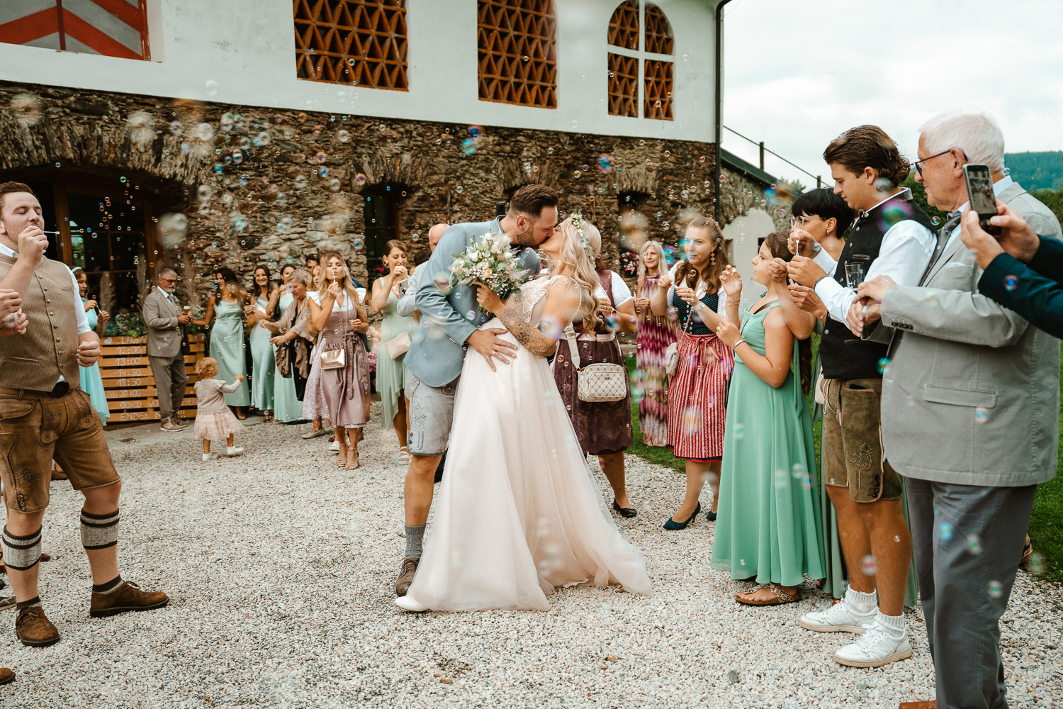 Ein Brautpaar küsst sich vor Lillis Feststadl in Feldkirchen nach Ihrer Hochzeit umringt von den Gästen. Das Foto wurde von Hochzeitsfotograf Valentino Zippo aus Kärnten aufgenommen.